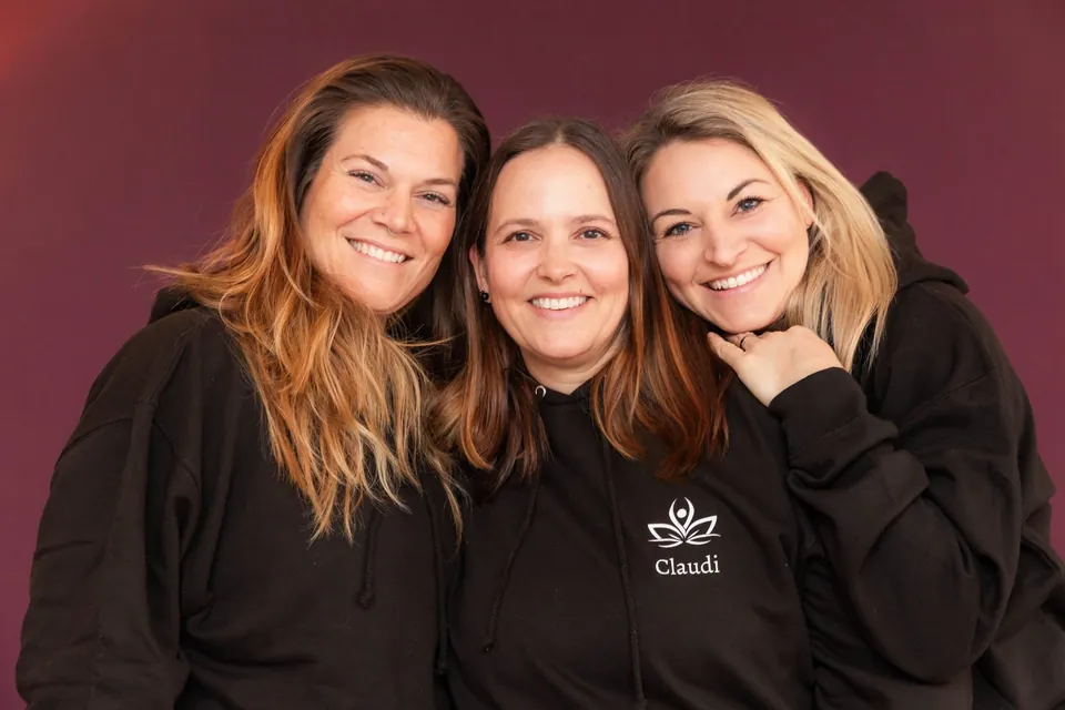 Die drei Kursleiterinnen des YogaStudio Pulheim lächeln gemeinsam in schwarzen Studio-Pullovern vor der beerenfarbenen Wand